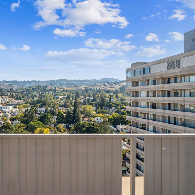 View from a balcony overlooking a residential area with many trees and houses, with hills in the background under a partly cloudy blue sky. A multi-story building is visible on the right side of the image.