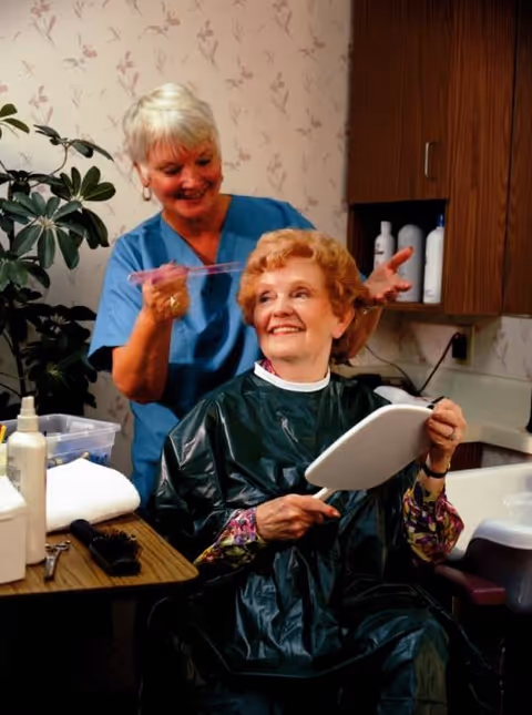 An elderly woman with curly hair sits in a salon chair wearing a black cape, holding a handheld mirror and smiling. A hairstylist in blue scrubs stands behind her, applying hair color with a brush. The setting appears to be a cozy salon area with floral wallpaper, a wooden cabinet with hair products, and a plant in the background.