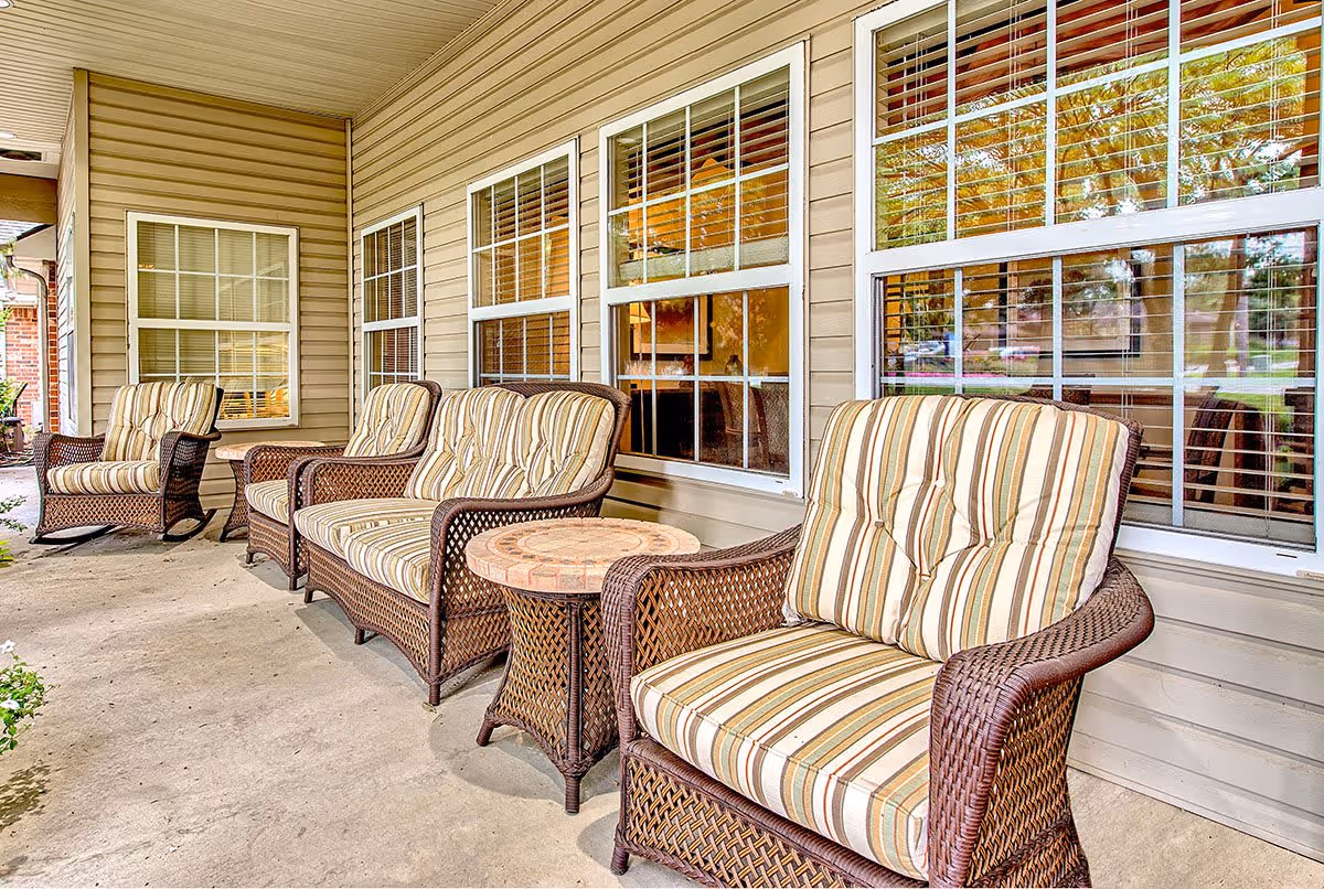 Outdoor patio area with four wicker chairs featuring striped cushions and a small round wicker table, situated in front of a building with beige siding and multiple windows with white frames and blinds.