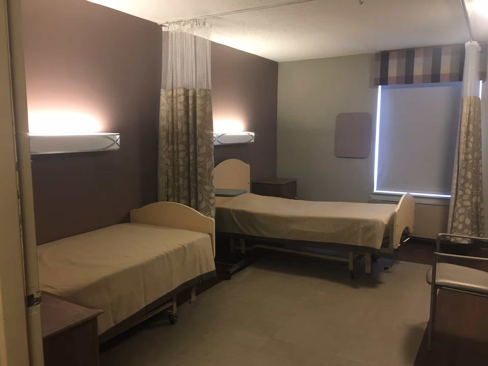 A room with two single beds separated by a privacy curtain. Each bed has a beige bedspread and a wooden headboard. The walls are painted in muted tones with wall-mounted lights above each bed. There is a window with a closed blind and a patterned valance, and a chair is visible near the window.