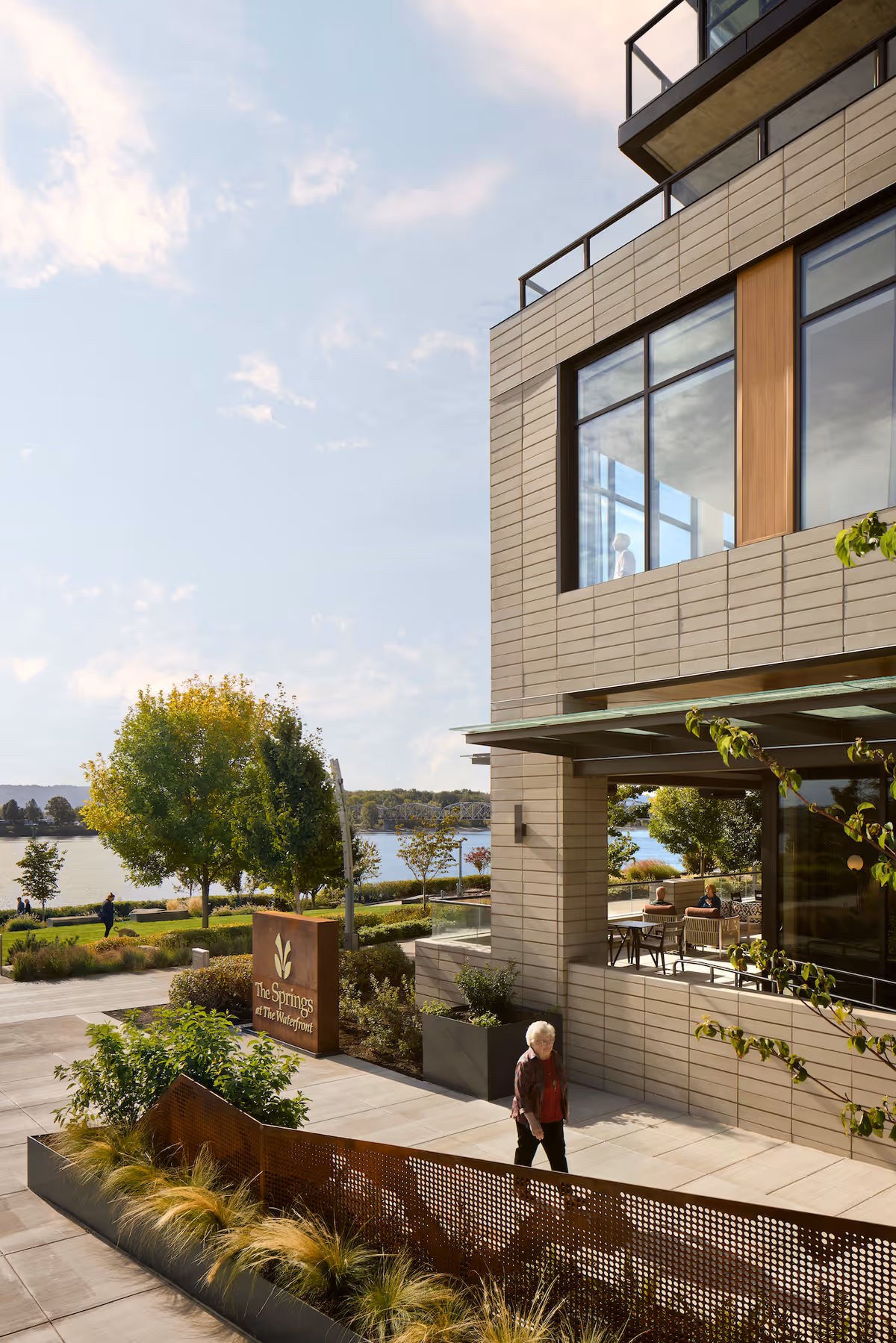 Exterior view of The Springs at The Waterfront senior living facility showing a modern building with large windows, outdoor seating area with people, landscaped walkways, trees, and a body of water in the background under a partly cloudy sky.