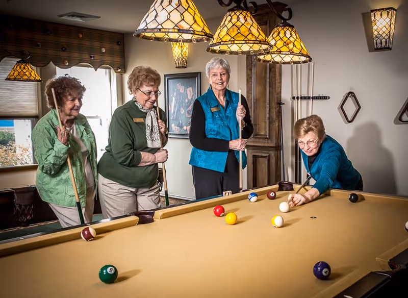 Four elderly women playing pool in a well-lit game room with hanging stained glass lamps above the pool table. One woman is taking a shot while the other three watch and hold pool cues. The room has a window with a valance, framed artwork on the wall, and a wooden cabinet in the background.