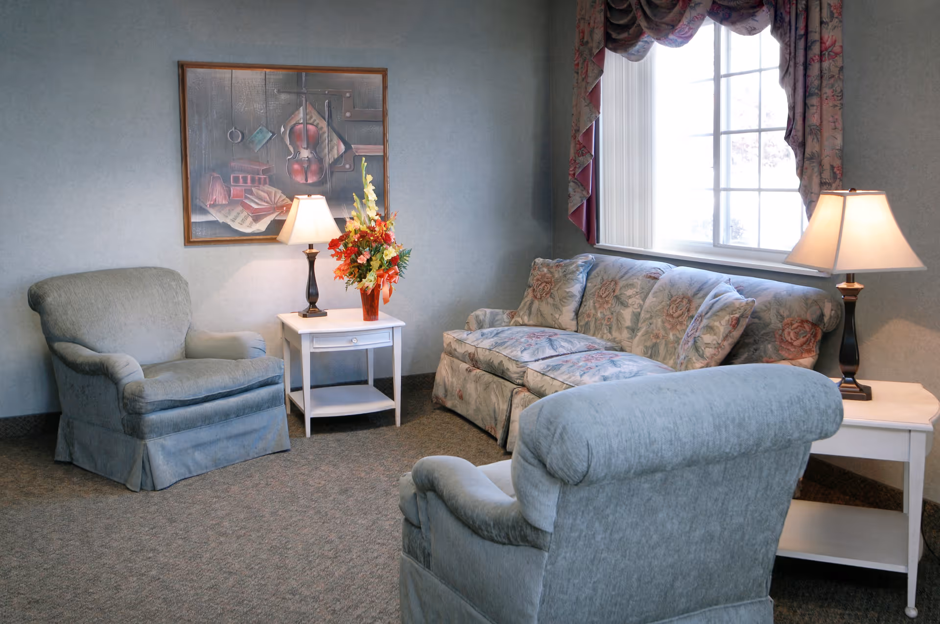 A cozy living room with two light blue upholstered armchairs and a floral patterned sofa. There are two white side tables, each with a lamp, and a vase with colorful flowers on one table. A large window with floral curtains lets in natural light, and a framed painting featuring musical instruments hangs on the wall.