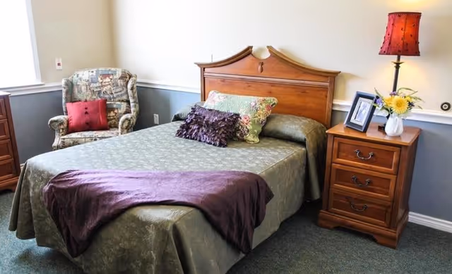 A bedroom with a wooden bed frame and a bed made with a green patterned bedspread, decorated with three pillows and a purple throw blanket. To the right of the bed is a wooden nightstand with three drawers, a red lampshade on a lamp, a framed photo, and a vase with yellow flowers. To the left of the bed is a patterned armchair with a red cushion. The walls are painted white and gray with a chair rail molding.