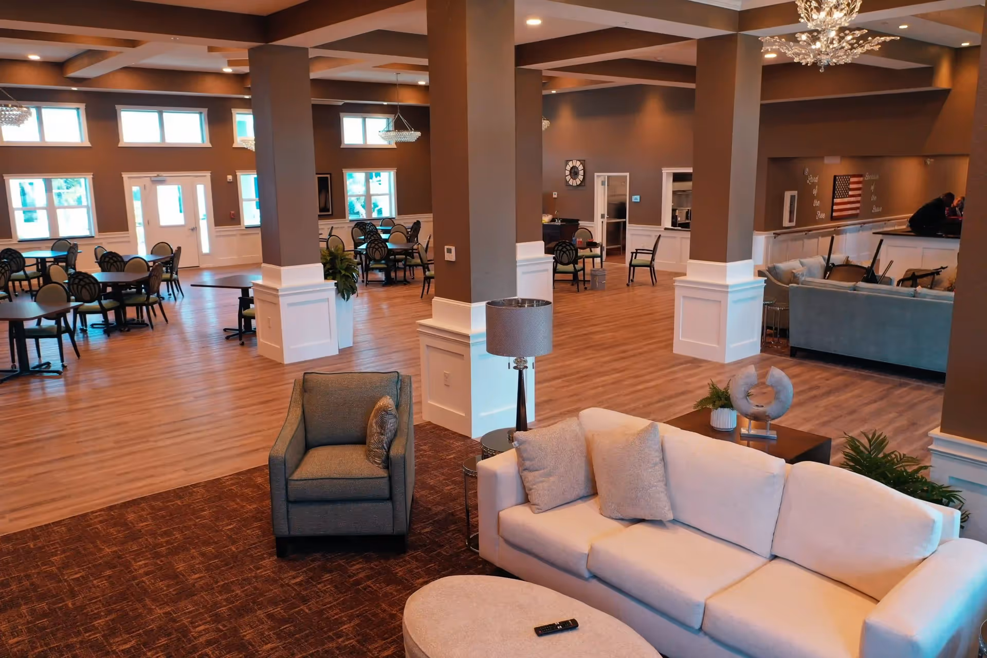 Spacious interior of a senior living facility featuring a seating area with a beige sofa, armchair, and a round ottoman in the foreground. The background shows multiple dining tables and chairs, large windows letting in natural light, and decorative columns with white paneling. The room has wood flooring and warm lighting with chandeliers.
