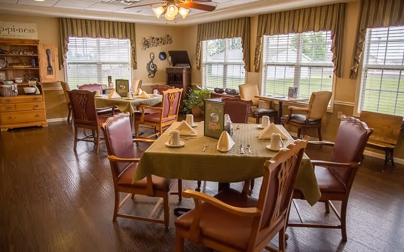 A cozy dining room with wooden floors and several tables set with napkins, cups, and utensils. The room has large windows with striped valances allowing natural light to fill the space. There are comfortable wooden chairs with cushioned seats around the tables, and decorative wall art including musical notes and a treble clef. A wooden cabinet with kitchenware is visible in the background.