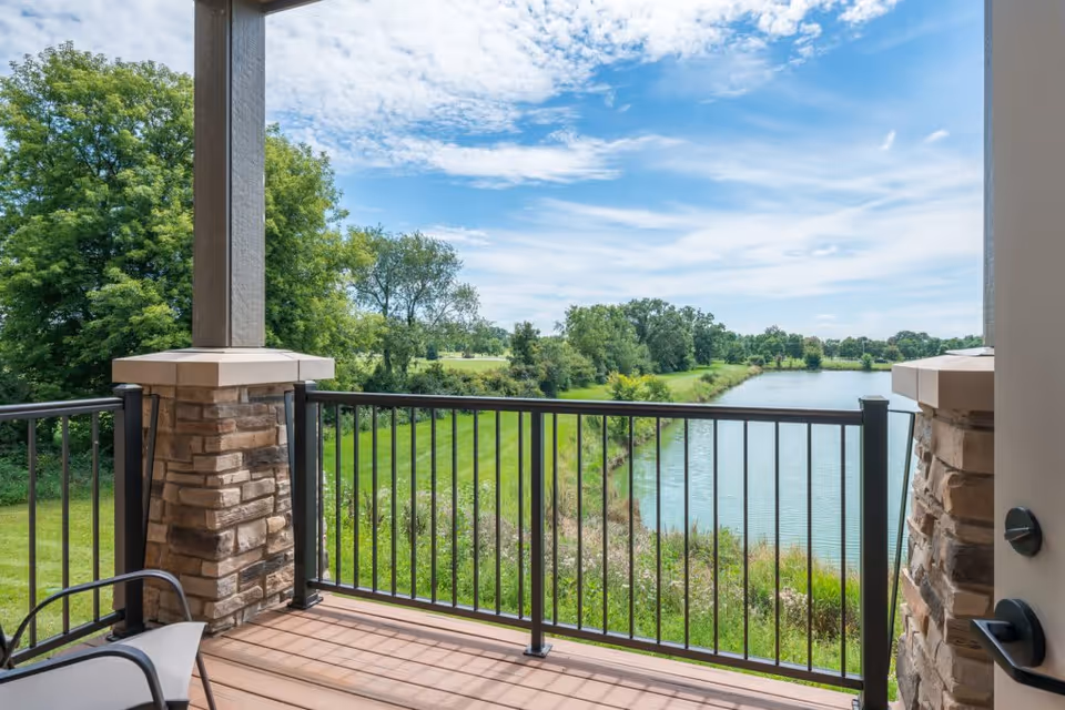 View from a covered balcony with stone pillars and black metal railing overlooking a green grassy area, trees, and a calm river under a partly cloudy blue sky.