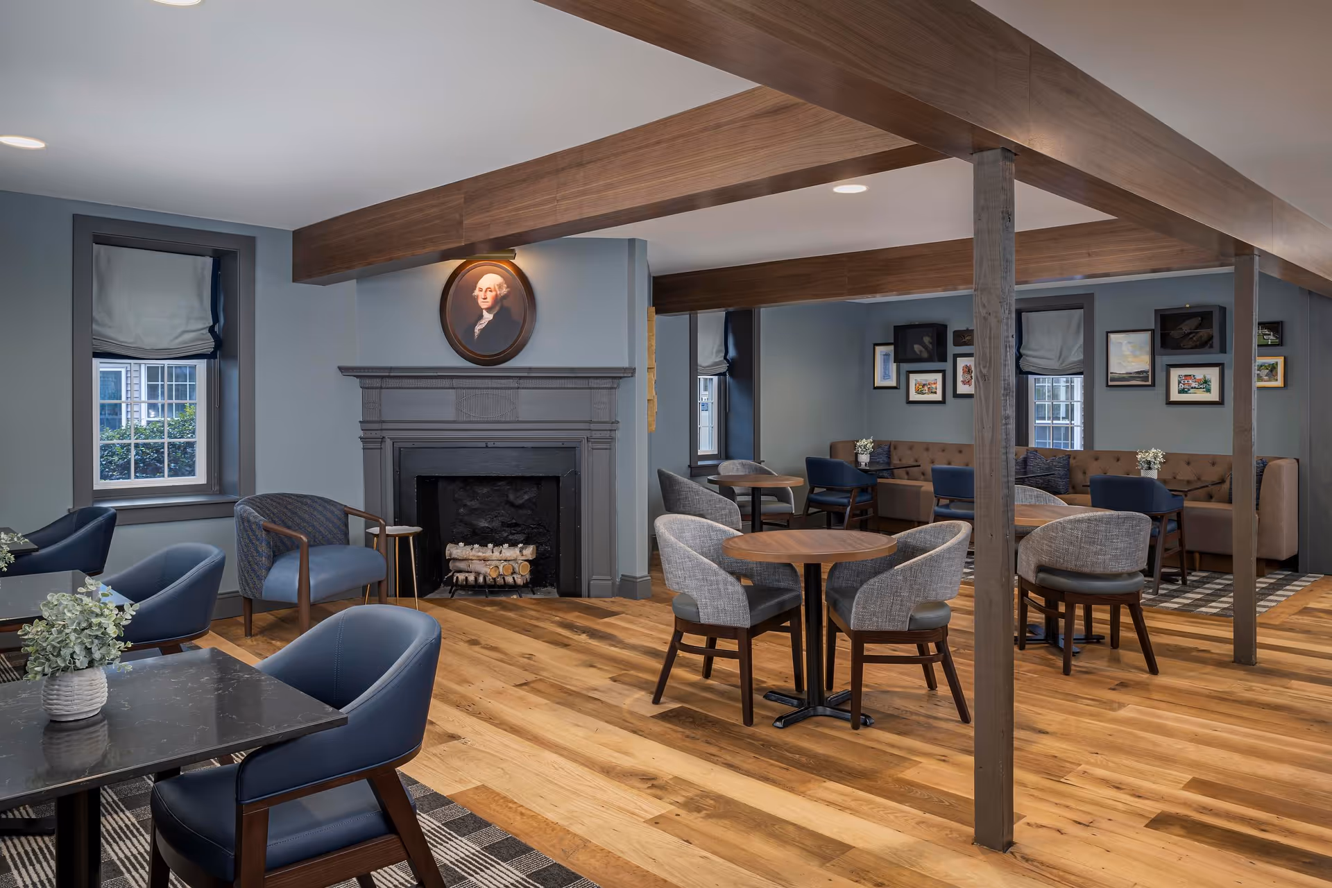 Comfortable communal dining and lounge area with round tables, upholstered chairs, a fireplace and a portrait on the wall.