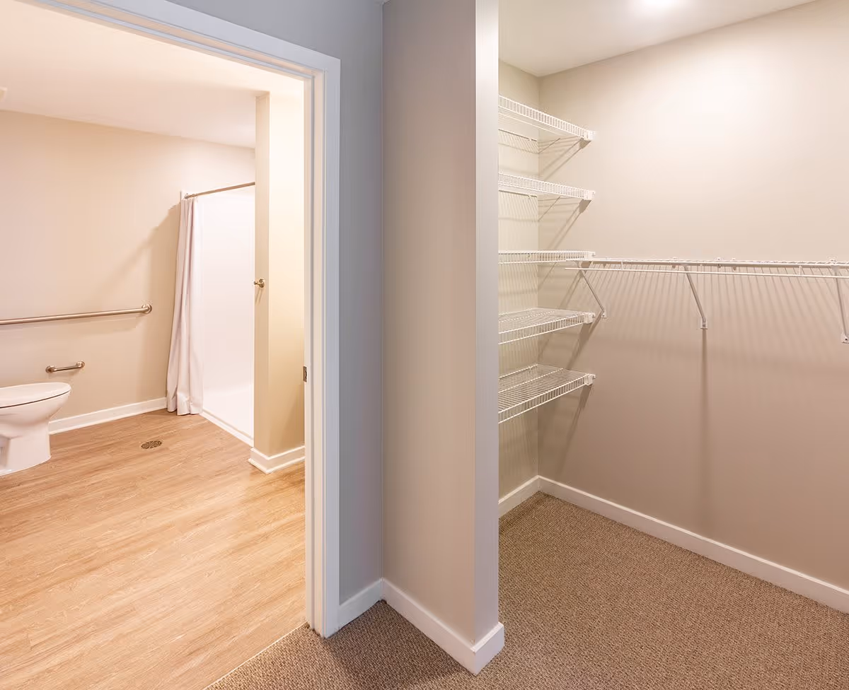 View of a bathroom with a toilet and a shower with a white curtain on the left side, and an empty walk-in closet with wire shelving on the right side, separated by a wall and doorway.