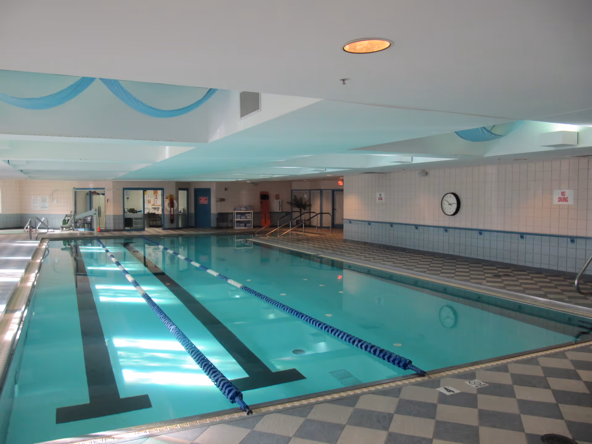 Indoor swimming pool with lane dividers in a tiled room featuring checkered floor tiles, a clock on the wall, and 'No Diving' signs.