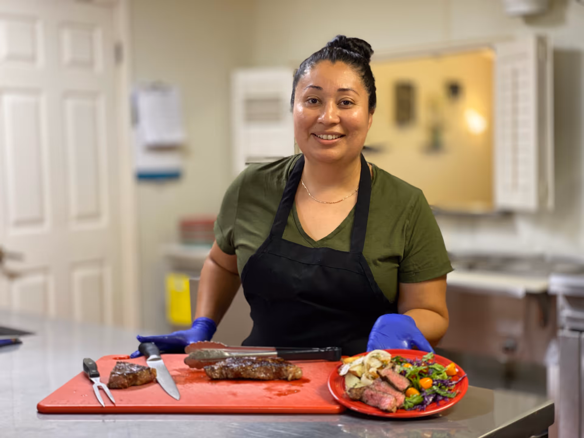 A woman wearing a green shirt, black apron, and blue gloves stands behind a kitchen counter with a red cutting board. On the board are a cooked steak, a knife, and a fork. She is holding a red plate with sliced steak, roasted potatoes, and a fresh salad. The background shows a kitchen area with white doors and a pass-through window.