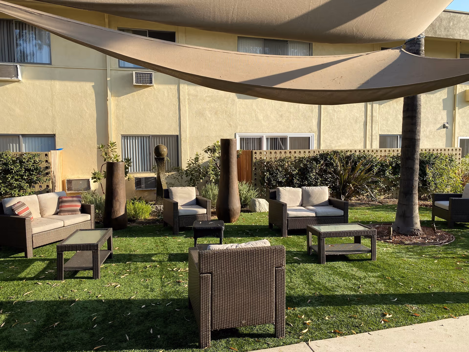 Outdoor seating area with wicker furniture including sofas and chairs with cushions arranged on a grassy lawn. There are two glass-top coffee tables and a large beige shade sail overhead. The background shows a beige building with windows and some plants along the wall.