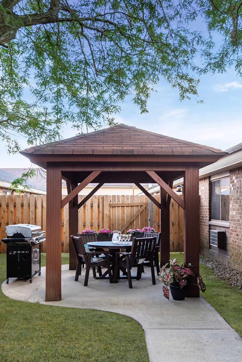 Outdoor gazebo with a wooden roof and four chairs around a round table set with glasses and a pitcher, located on a concrete patio in a fenced backyard. A barbecue grill is positioned nearby, and there are potted plants and a tree overhead.