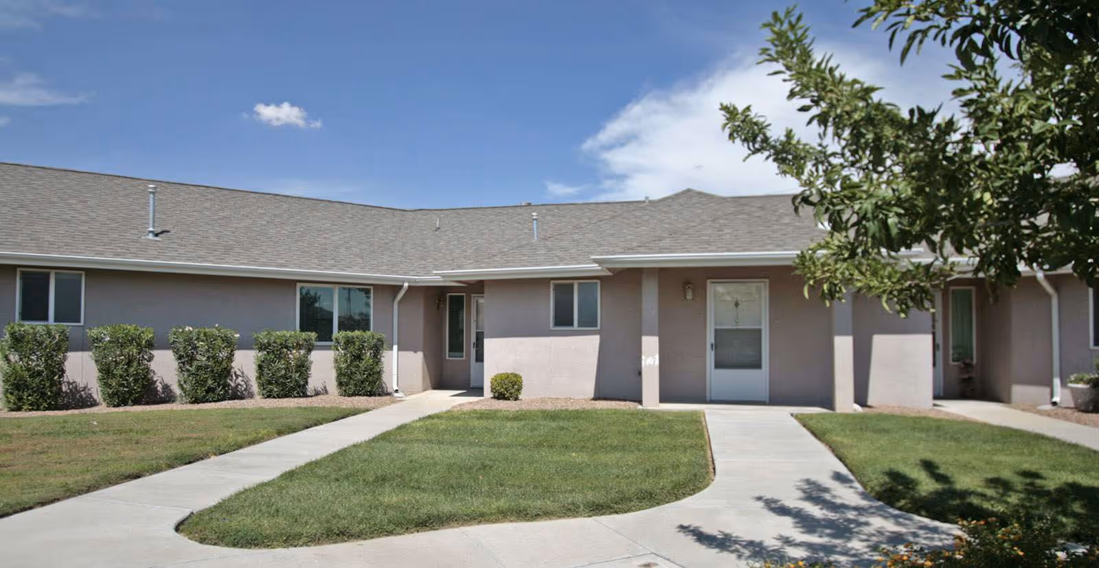 Single-story beige senior living building front with entry doors, concrete walkways, trimmed shrubs, lawn and a tree under a blue sky.
