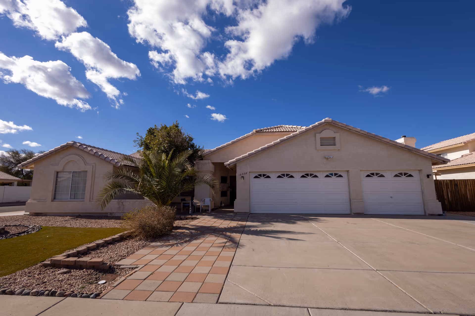 Single-story stucco house with a three-car garage, tiled roof, palm tree, and paved driveway under a blue sky.