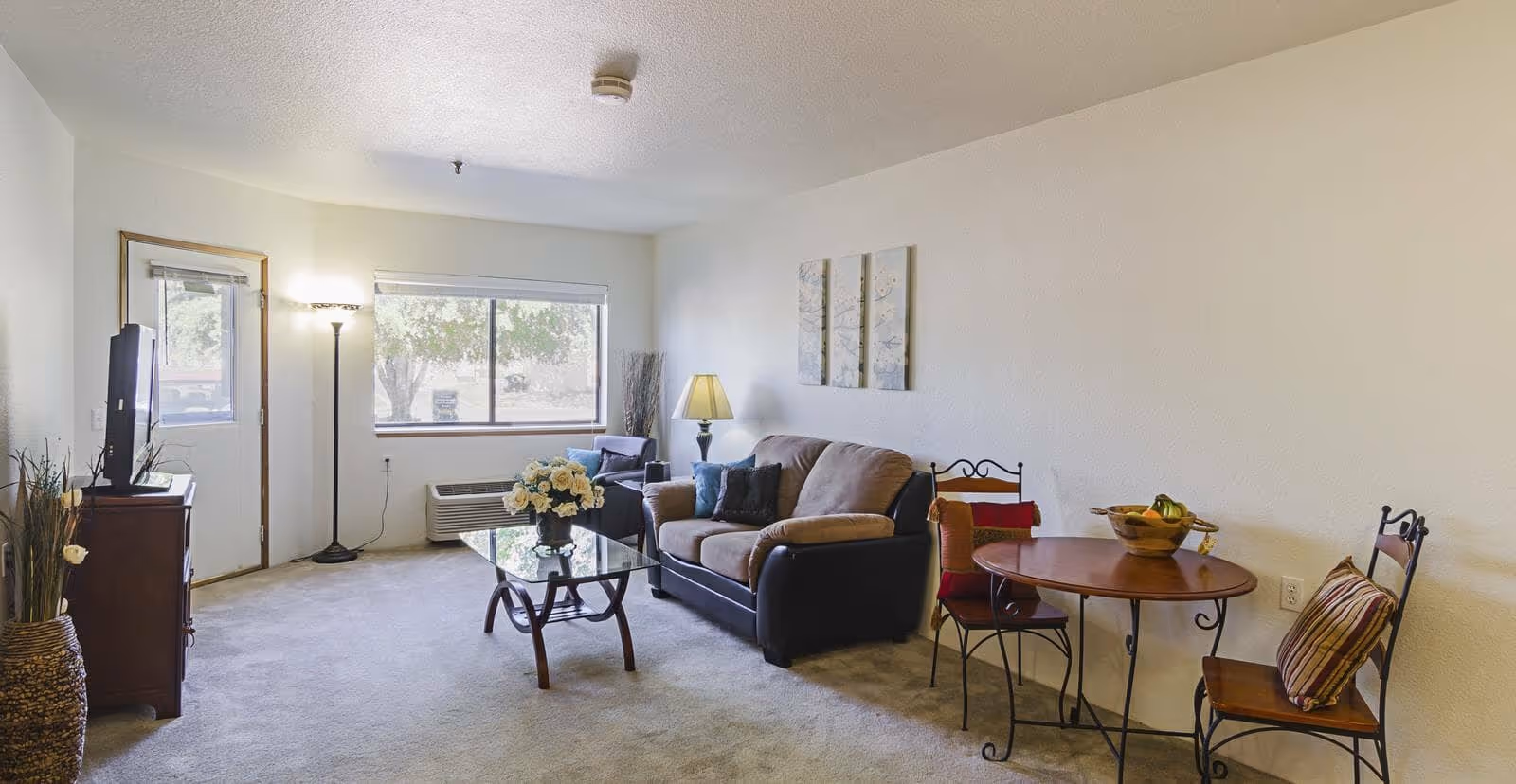 Bright furnished living room with a sofa, glass coffee table, TV, and a small round dining table by a window.