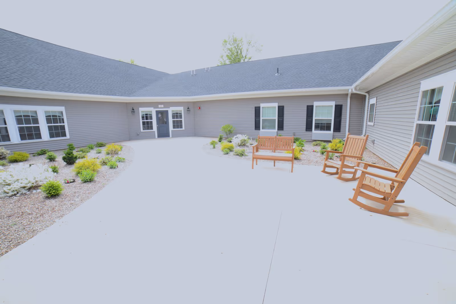 A paved courtyard at a single-story memory care building with landscaped beds and wooden rocking chairs and a bench.