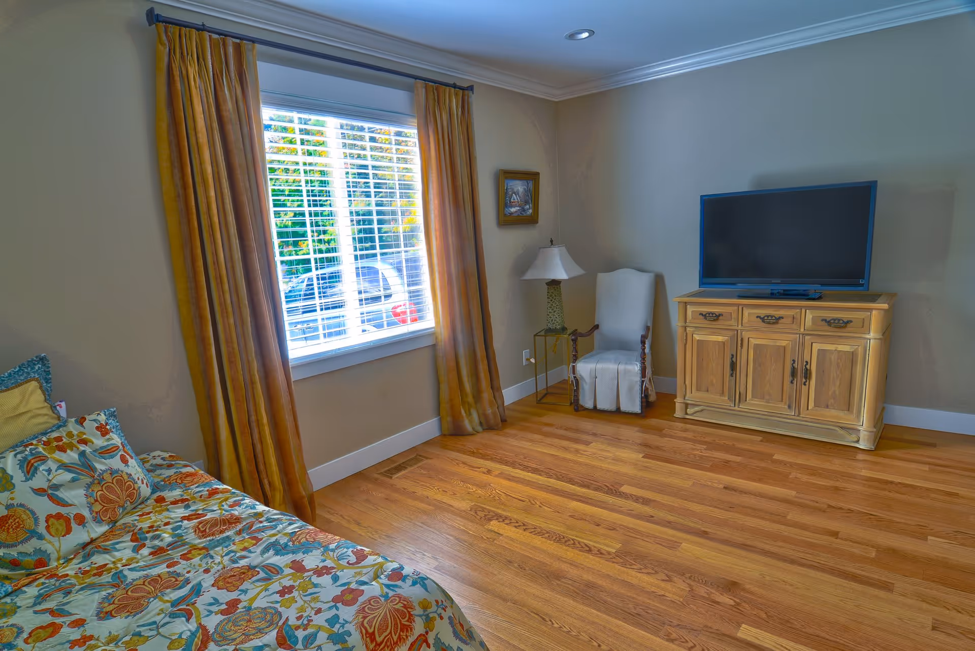 A cozy room with a floral patterned bedspread on a bed, a window with yellow curtains, a white armchair next to a small side table with a lamp, and a wooden cabinet with a flat-screen TV on top. The room has wooden flooring and beige walls.
