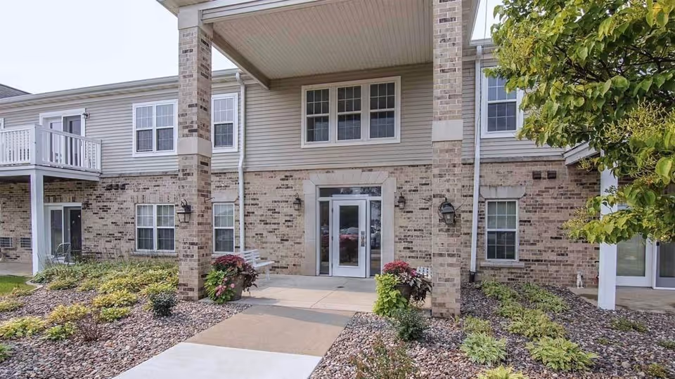 Front entrance of a two-story brick-and-siding apartment building with a covered entry, walkway, and landscaped beds.