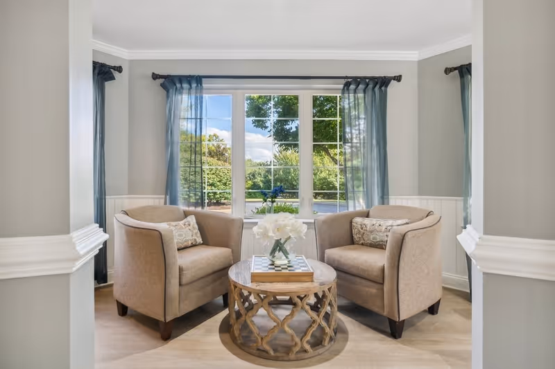 A cozy sitting area with two beige armchairs facing each other, separated by a round wooden coffee table with a decorative lattice base. On the table is a vase with white flowers and a chessboard. Behind the chairs is a large window with blue sheer curtains, showing a green outdoor landscape with trees and bushes.