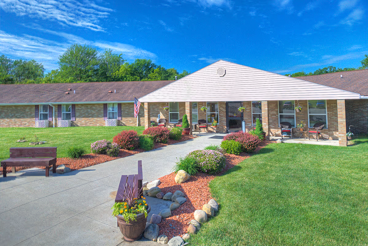 Exterior view of Dixon Healthcare Center showing a single-story brick building with a covered entrance. There is a paved walkway leading to the entrance, surrounded by well-maintained green lawns, flower beds with shrubs and rocks, and outdoor seating. The sky is clear and blue with some clouds.