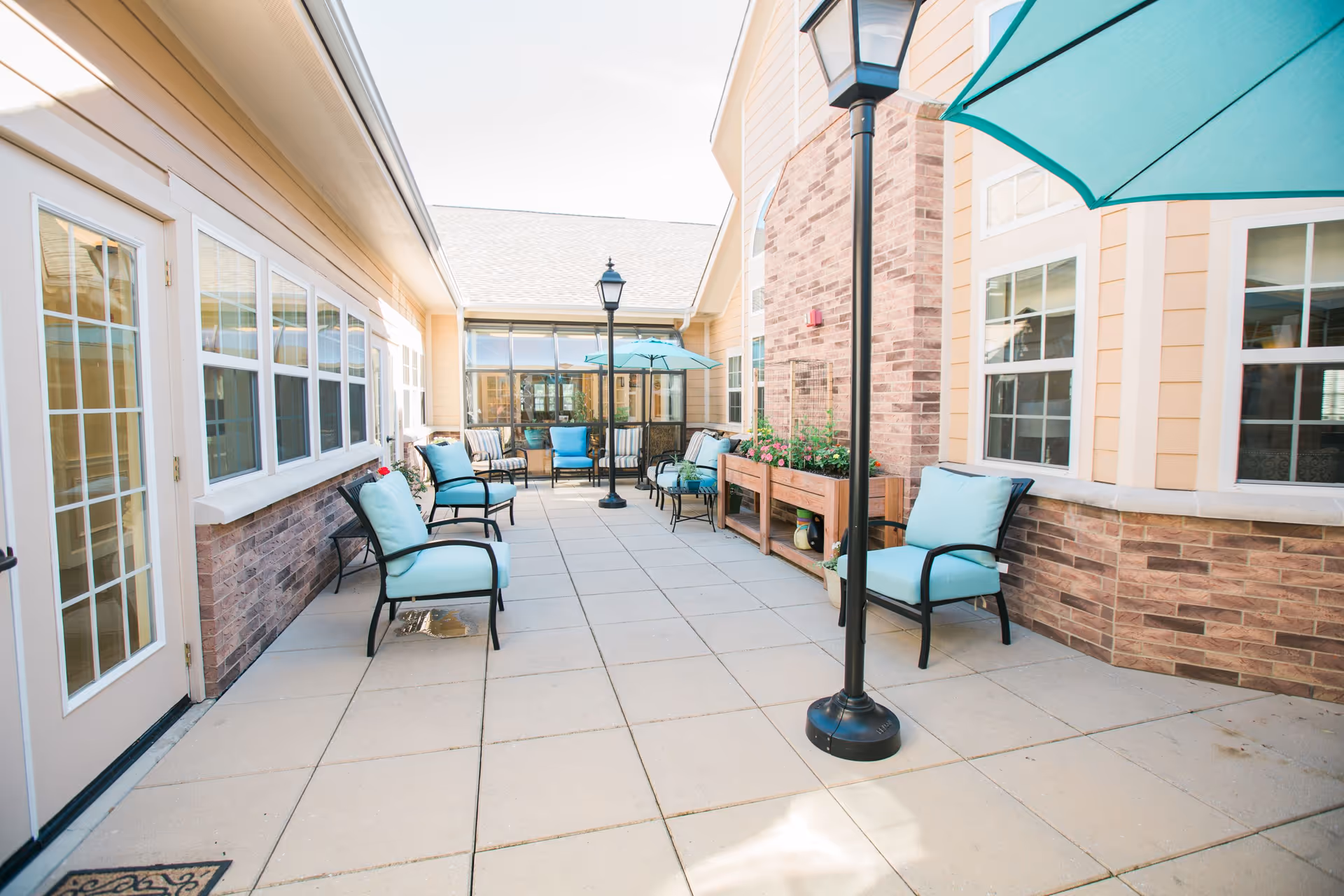 Outdoor patio area at a senior living facility with several cushioned chairs arranged along the walls, a large teal umbrella providing shade, two black lamp posts, and planter boxes with flowers. The area is surrounded by windows and brick walls.