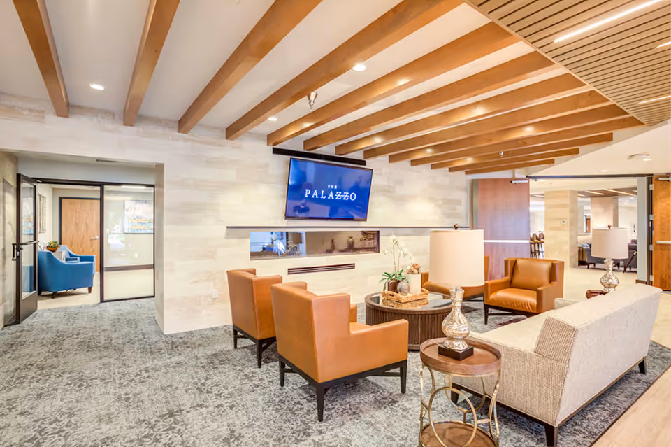 A modern and spacious living room area in The Palazzo facility featuring a beige sofa, two brown leather armchairs, a round coffee table with decorative items, and two side tables with lamps. The room has a carpeted floor, wooden ceiling beams, and a wall-mounted TV displaying 'The Palazzo'. Glass doors lead to another room with a blue armchair.