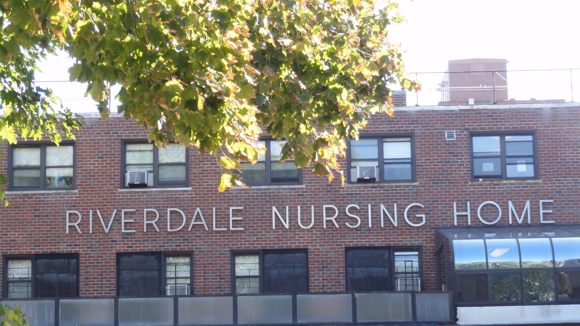 Exterior view of a brick building with multiple windows and a sign that reads 'RIVERDALE NURSING HOME' partially obscured by tree branches with green leaves.