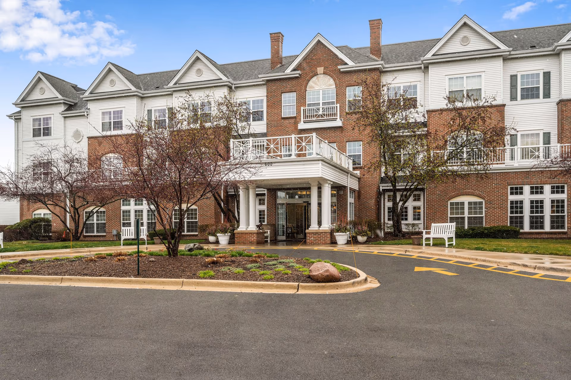 Front exterior view of a three-story senior living facility building with a combination of red brick and white siding. The entrance features a covered portico with white columns and potted plants. There are trees and landscaping in front, a bench on the right side, and a driveway with yellow directional arrows leading to the entrance. The sky is partly cloudy.