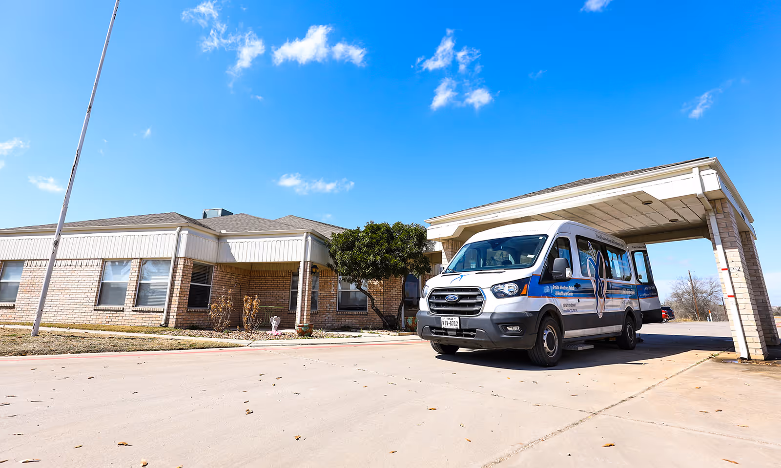Exterior view of Prairie Meadows Rehabilitation and Healthcare Center showing a single-story brick building with multiple windows and a covered entrance. A white and blue shuttle van is parked under the covered entrance. The sky is clear with a few clouds.
