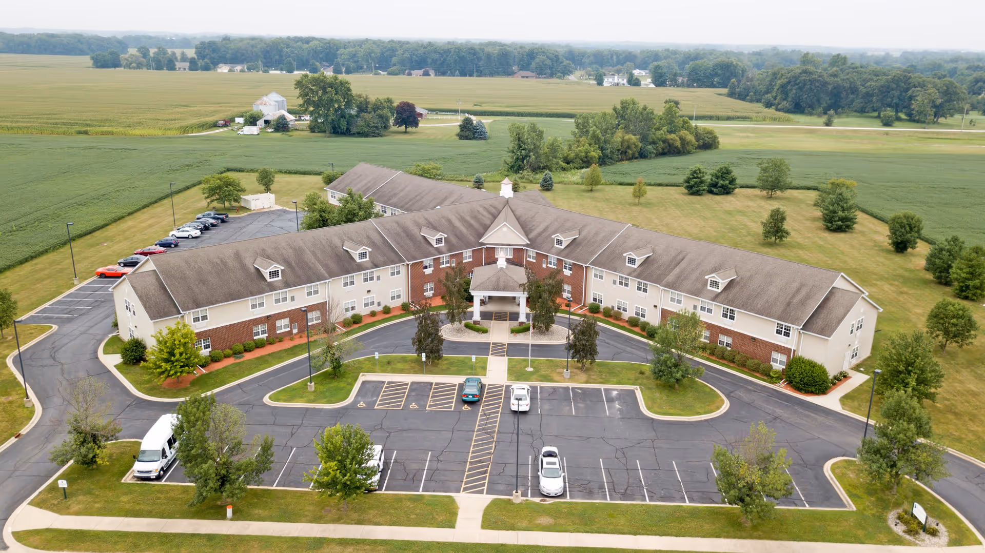 Aerial view of a large senior living facility building with a U-shaped layout surrounded by green lawns and trees. The building has a combination of brick and beige siding with multiple windows and a central entrance with a covered porch. There is a parking lot in front with several cars parked and a van on the left side. The facility is set in a rural area with open fields and farmland in the background.