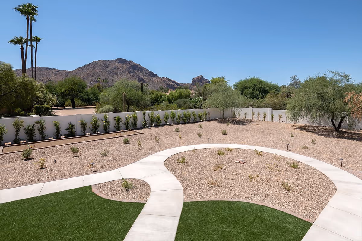 Outdoor garden area with a curved concrete pathway, gravel landscaping with small plants, green grass patches, and a white fence. In the background, there are trees, tall palm trees, and a mountain under a clear blue sky.