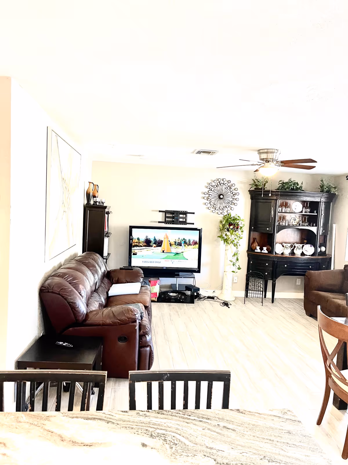 A bright living room with a brown leather sofa on the left, a flat-screen TV mounted on the wall in the center, and a dark wooden cabinet with decorative plates and plants on the right. A ceiling fan with wooden blades is mounted on the white ceiling. Light-colored wood flooring covers the room, and a dining table with chairs is partially visible in the foreground.