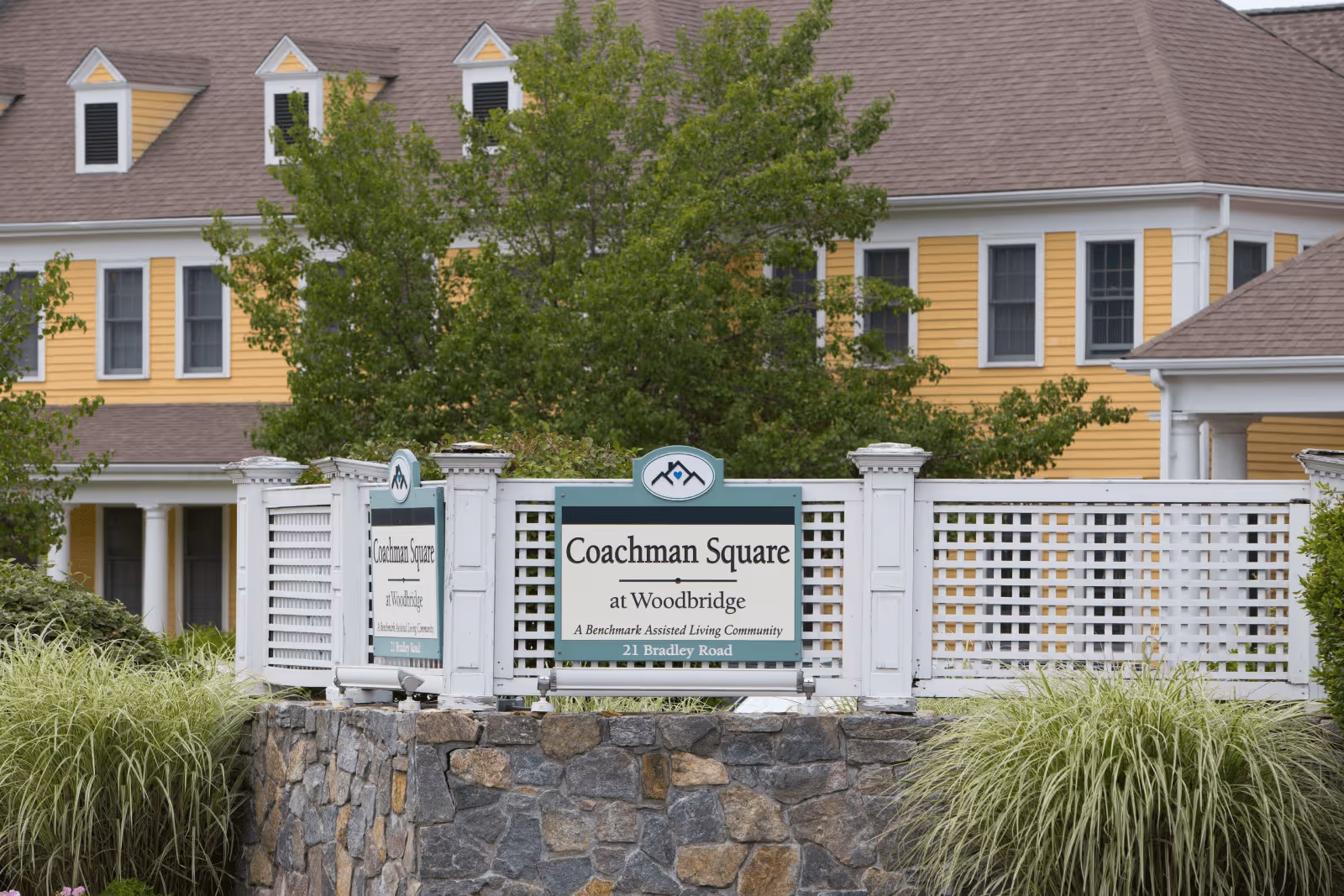 Exterior view of Coachman Square at Woodbridge, an assisted living community, showing a yellow building with white trim behind a white lattice fence and stone wall with a sign displaying the community's name and address.