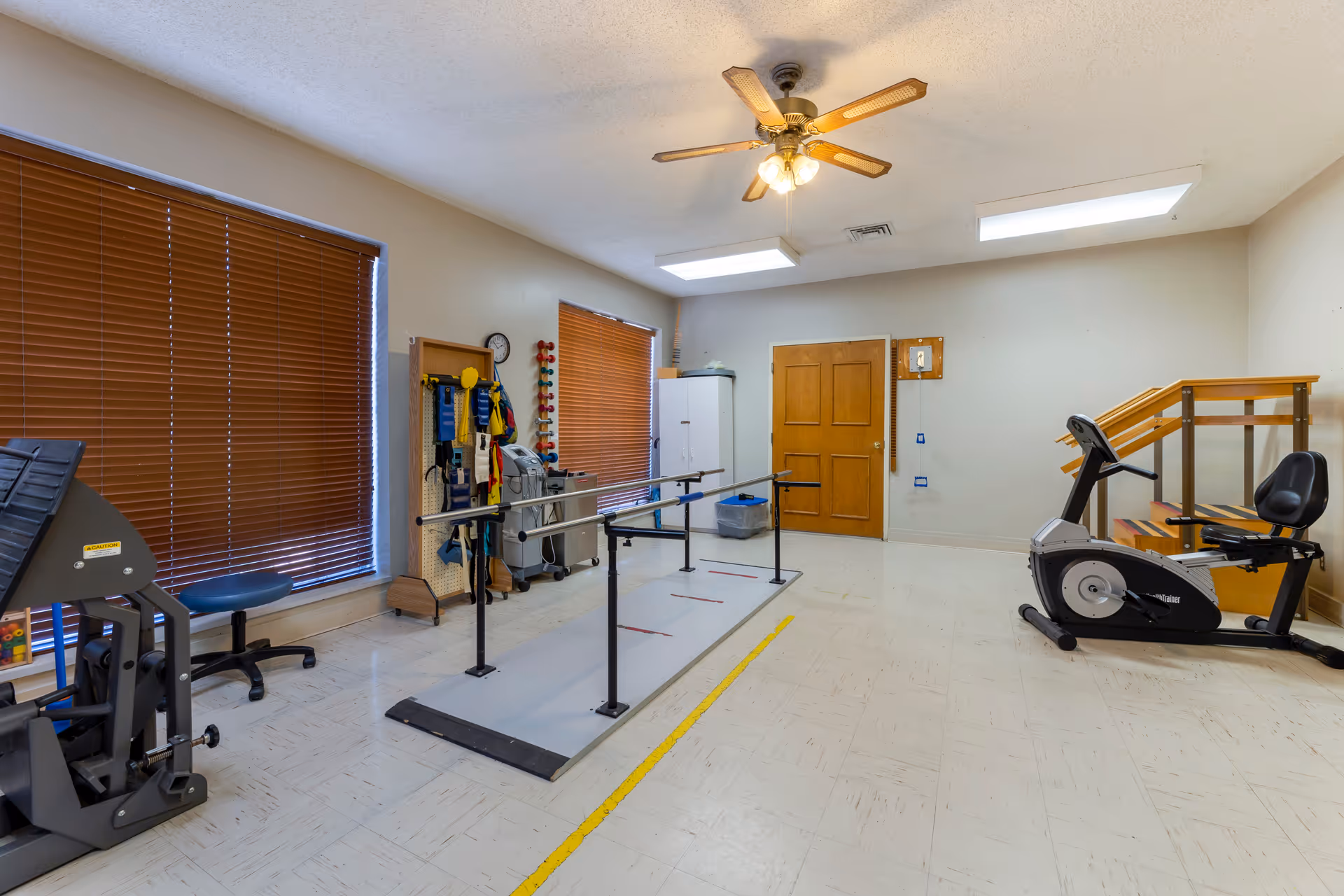 A rehabilitation room with exercise equipment including parallel bars for walking practice, a recumbent exercise bike, and a set of stairs with handrails. The room has two windows with brown blinds, a ceiling fan with lights, and a white cabinet against the wall.