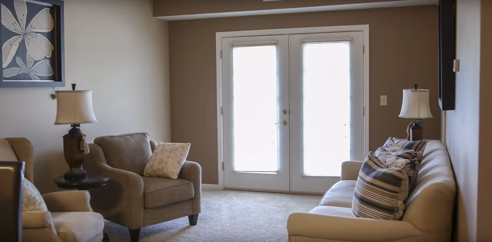 A cozy living room with a beige sofa adorned with striped and floral pillows, two armchairs with decorative cushions, two table lamps on small round tables, and a large framed floral artwork on the wall. French doors with blinds let in bright natural light.