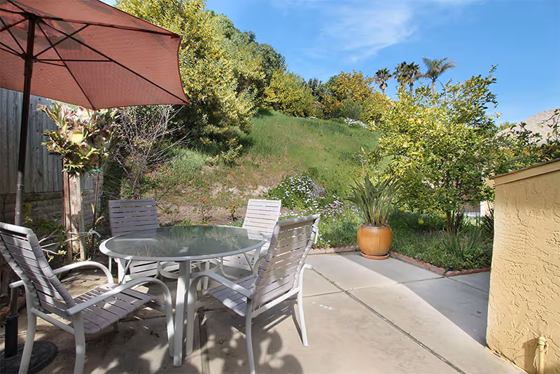 Outdoor patio area with a round glass table and four white chairs under a large pink umbrella. The patio is surrounded by green plants, trees, and a grassy hill under a clear blue sky.