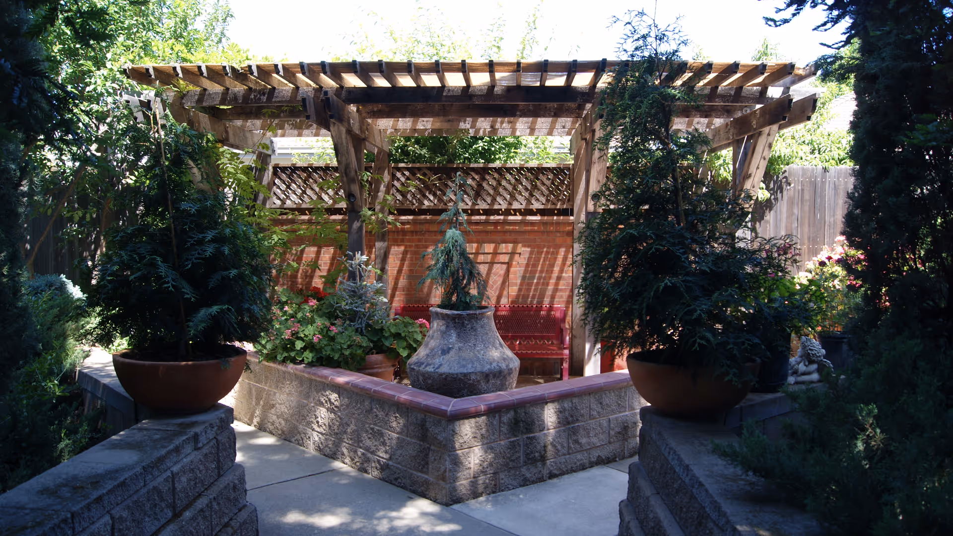 Sunlit courtyard with a wooden pergola over a central stone planter, potted evergreens, and a bench.
