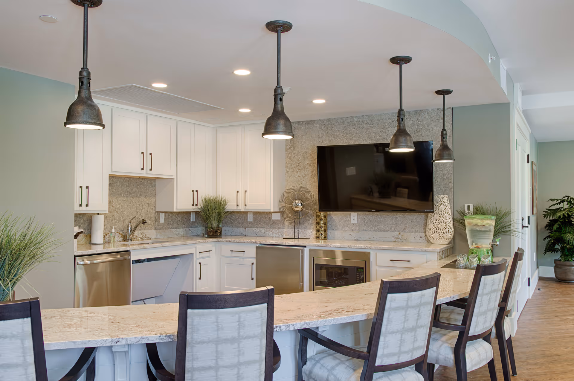 Modern kitchen area with white cabinets, granite countertops, and stainless steel appliances including a dishwasher and microwave. Four upholstered chairs are placed along a curved countertop bar. Three pendant lights hang from the ceiling above the bar. A flat-screen TV is mounted on the wall above the countertop, and decorative plants and items are placed around the kitchen.