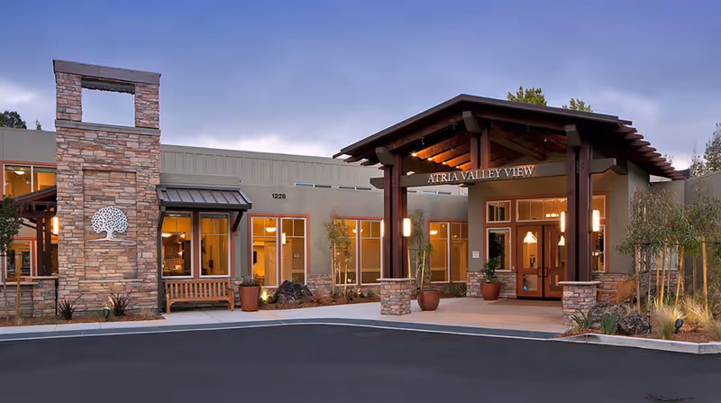 Exterior view of Atria Valley View senior living facility at dusk, showing the entrance with a wooden pergola, stone pillars, large windows, and a paved driveway with landscaping.