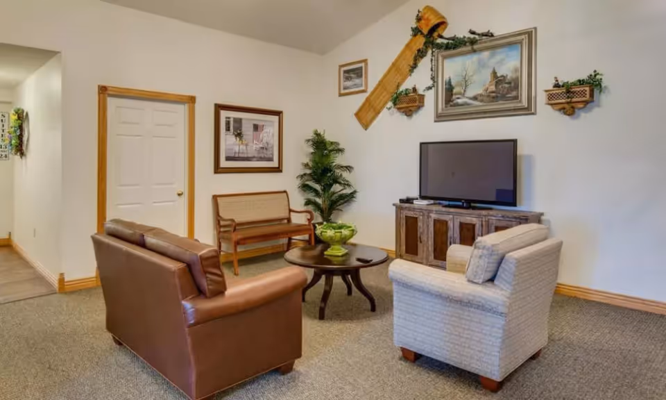 A cozy living room area with a brown leather loveseat, a beige armchair, and a wooden bench against the wall. A round wooden coffee table with a green decorative bowl is in the center. A flat-screen TV sits on a rustic wooden cabinet. The walls are decorated with framed paintings, a wooden sled, and small shelves with plants. The room has beige carpet and light-colored walls with wooden trim.