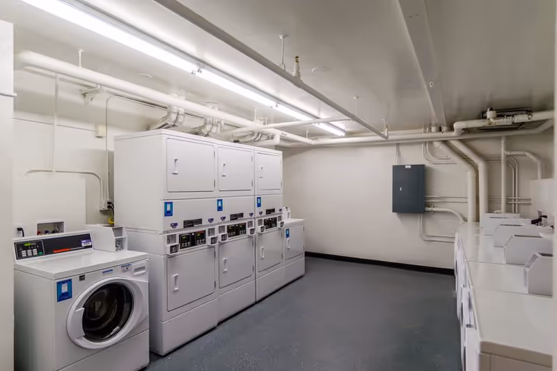 A clean communal laundry room with stacked dryers and multiple washing machines under fluorescent lighting.