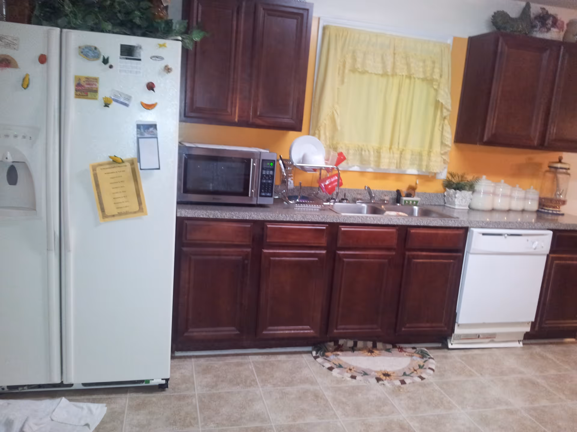 A kitchen with dark wooden cabinets, a white refrigerator with magnets and papers attached, a microwave on the countertop, a dish rack with dishes, a double sink, a dishwasher, and a window with yellow curtains. The walls are painted yellow and the floor is tiled.