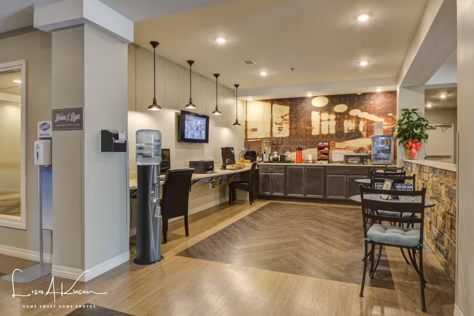 A cozy and well-lit common area with a coffee and snack station along the back wall, featuring cabinets, a microwave, coffee machines, and a small refrigerator. There are several black chairs and small round tables with cushions on the right side. On the left side, a water cooler and a computer workstation with chairs are visible. The floor has a wood pattern, and the walls are decorated with a large mural and pendant lights hanging from the ceiling.