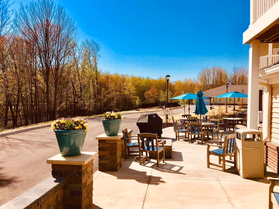 Outdoor patio area with tables, chairs, umbrellas, planters and a grill next to a senior living building with trees in the background.