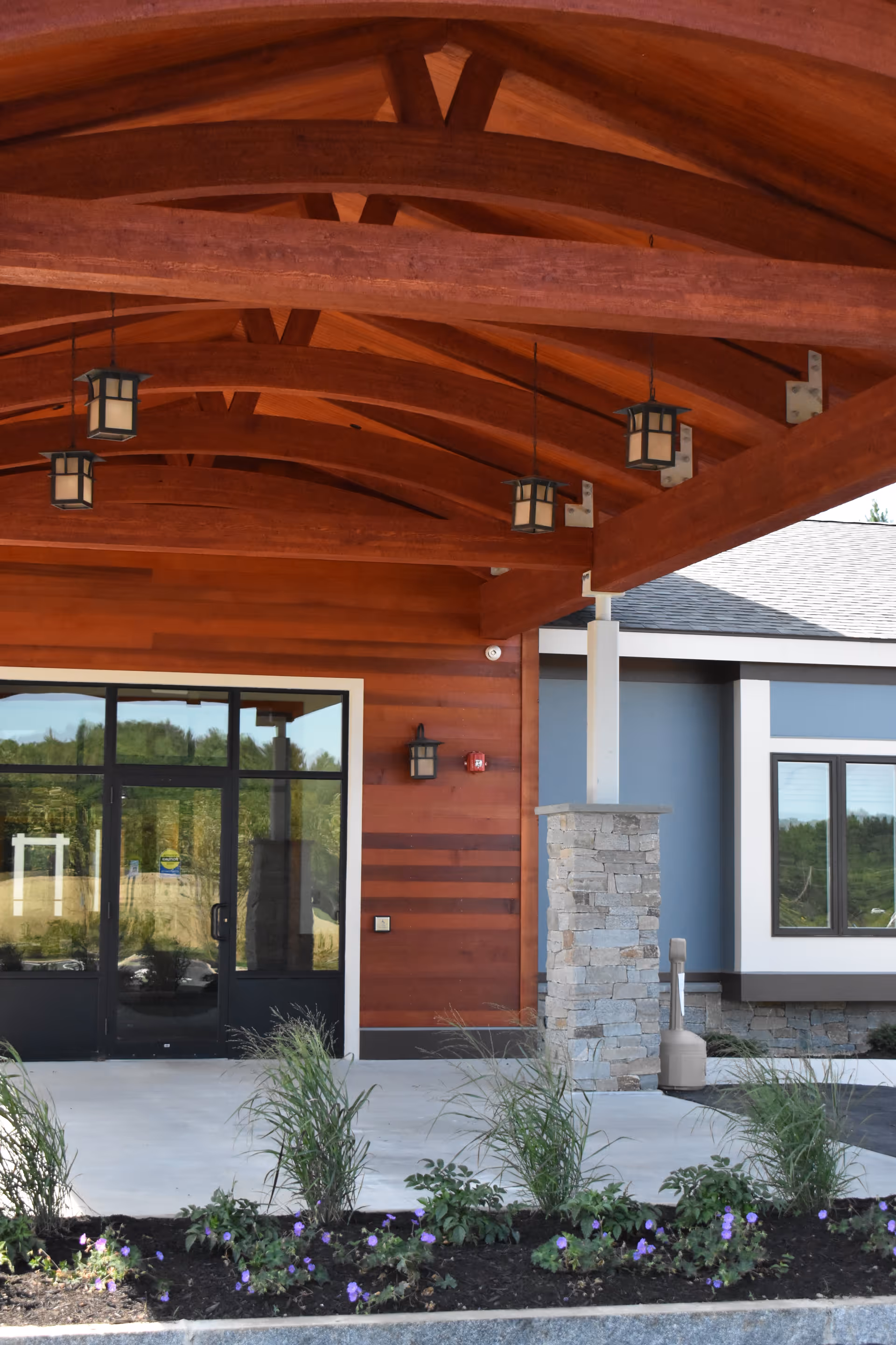 Entrance of a senior living facility with a wooden arched roof supported by stone pillars, glass double doors, and a landscaped flower bed with green plants and purple flowers in front.