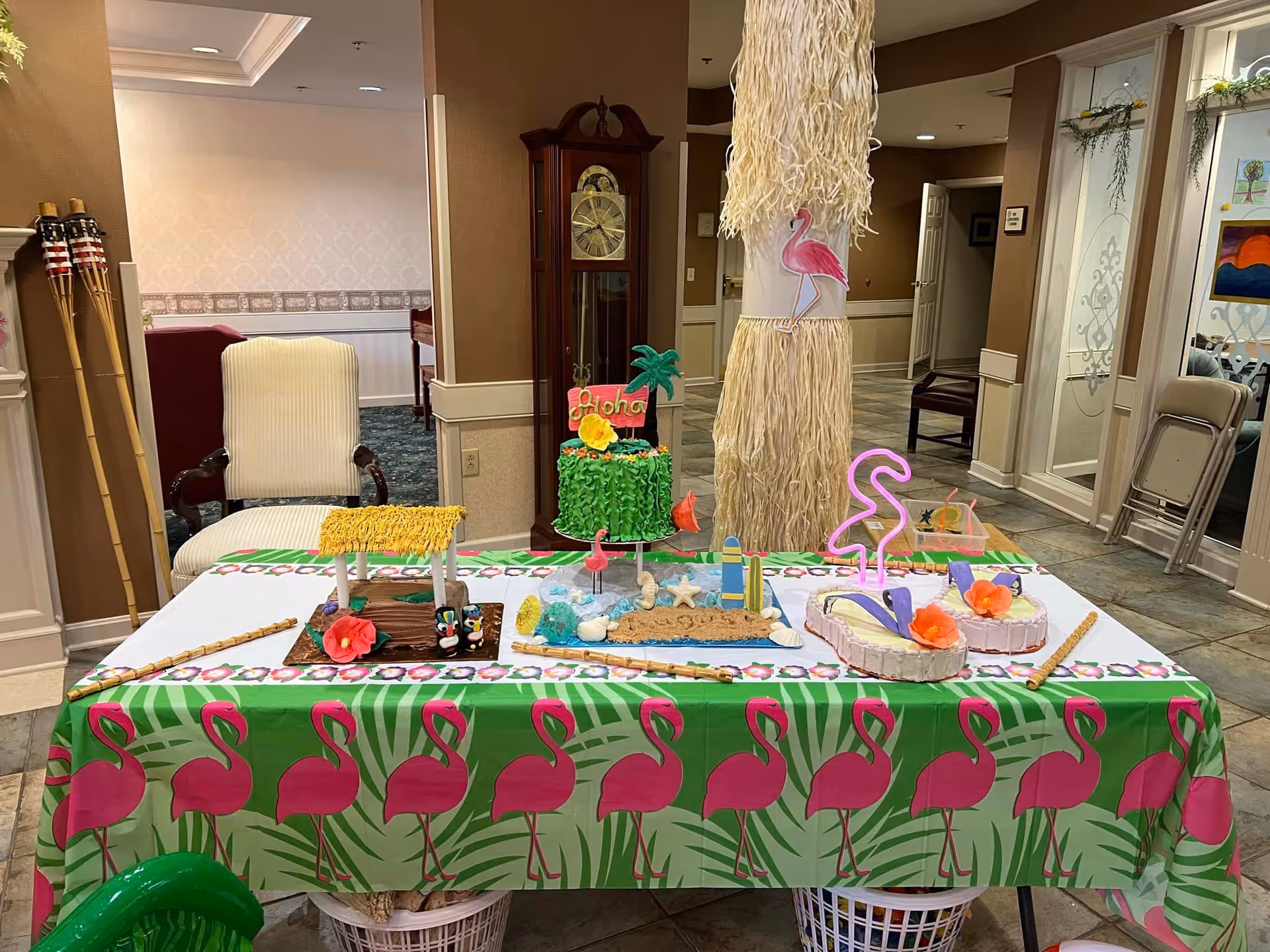 A decorated table with a green tablecloth featuring pink flamingos, displaying tropical-themed cakes and decorations including a cake with 'Aloha' written on it, a small hut cake, and flamingo figures. The background shows an indoor area with chairs, a grandfather clock, tiki torches, and a column wrapped with grass skirt material and a flamingo cutout.