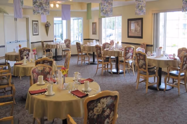 A dining room with round tables covered in yellow tablecloths, each set with cups, glasses, plates, and red napkins. The chairs have floral upholstery. The room is well-lit with large windows letting in natural light and decorative hanging fabric pieces from the ceiling.