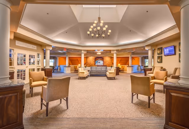 Spacious and well-lit senior living facility common area with beige carpet, multiple seating arrangements including armchairs and sofas, white columns, a chandelier hanging from a high ceiling, and a sign indicating the Muriel Major Library on the left side.
