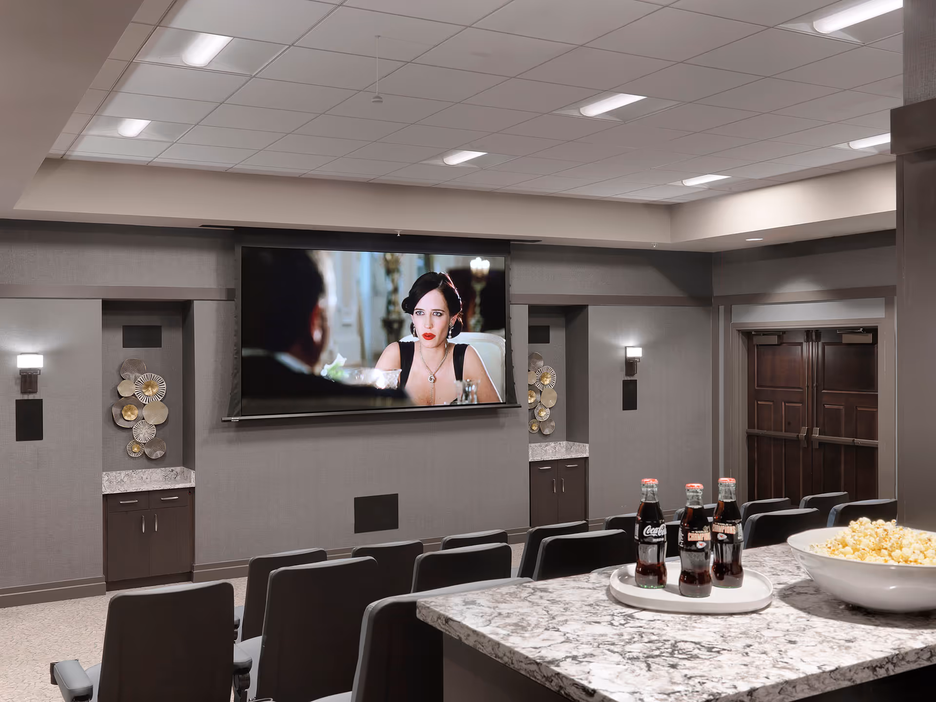 A senior living facility movie theater room with rows of black chairs facing a large screen showing a woman in a black dress. In the foreground, there is a marble countertop with a bowl of popcorn and a tray holding four bottles of Coca-Cola. The room has gray walls, decorative wall art, and double wooden doors at the back.